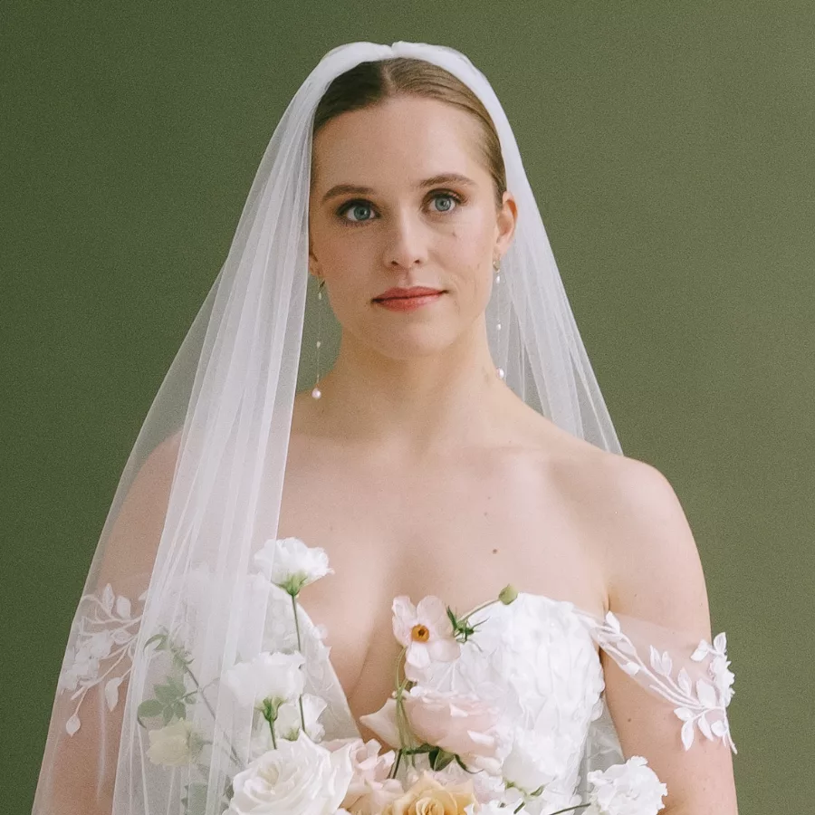 Long drop pearl bridal earrings being worn by a bridal model, wearing a veil and has her hair back in a bun. She wears a bridal gown with a straps that drape below her shoulders and she holds a bridal bouquet with peach and white flowers