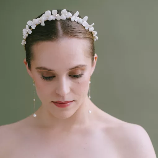 ivory white floral bridal crown with pearls being worn by a bride who wears her hair in a sleek centre parted bun. She has blonde hair and wears long chain pearl earrings against a green backdrop.