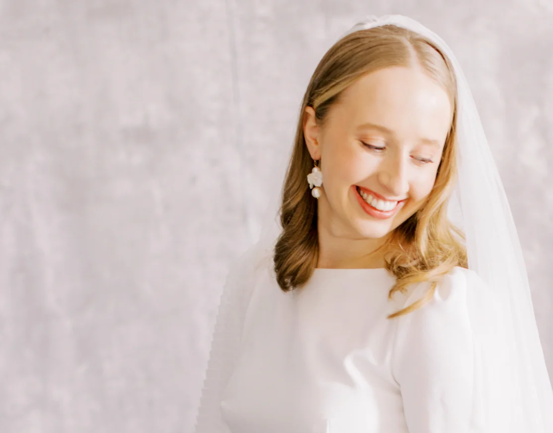 bride looking down smiling. She is stood in front of a white fabric background. She wears a white high neck wedding dress, with veil and earrings. 5 tips for stress-free wedding accessory and jewellery shopping