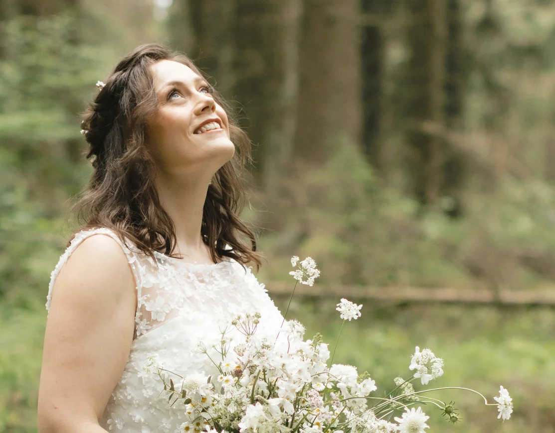 Woman looking up in a forest, wearing a wedding dress and holding a bridal bouquet to reflect the topic Elevate Your 2025 Woodland Wedding with These Hairpieces